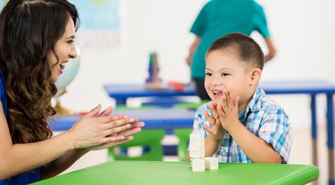 A teacher and student with Down Syndrome clap together at a desk.