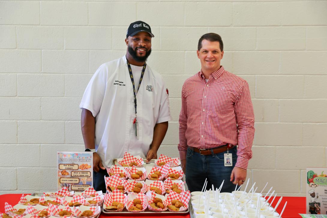 Two men stand behind a display of sample fair foods