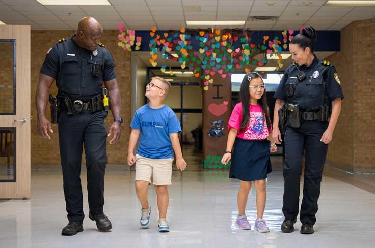School resource officers walking smiling at two young students as they walk down the hall together.