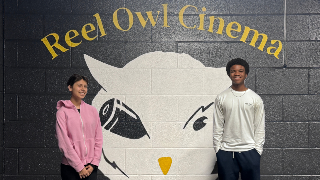 One female and one male student stand in front of a wall with the Reel Owl Cinema logo 
