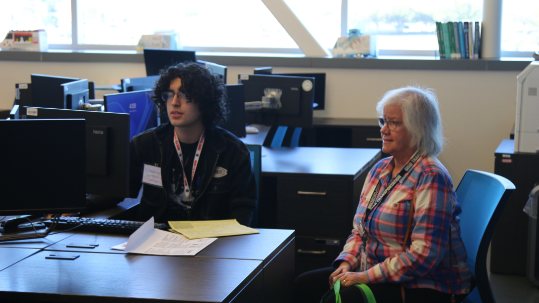 A male student works at a computer as an adult female sits next to him