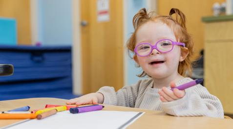 A young student with Down Syndrome smiles for the camera while she uses crayons.